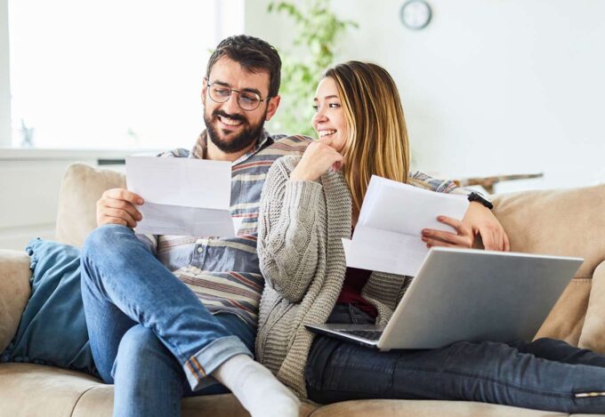 Couple at home on the couch looks at paperwork and laptop to consider the pros and cons of refinancing their home.