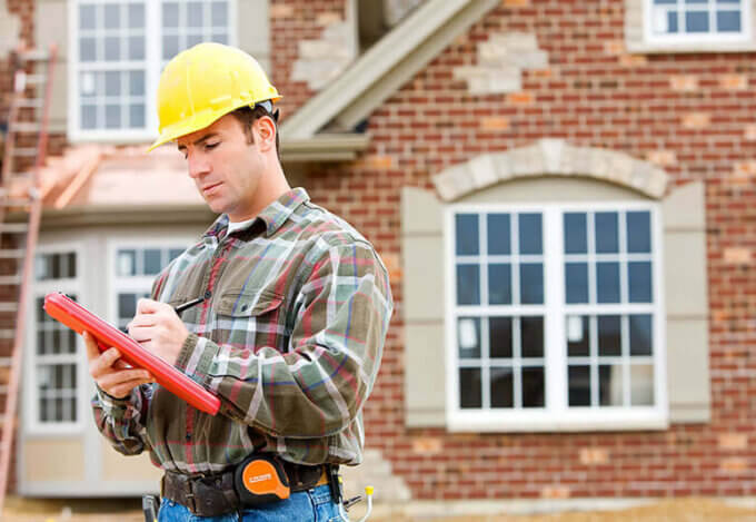 Home inspector in a hard hat reviewing a home inspection checklist in front of a brick house under evaluation